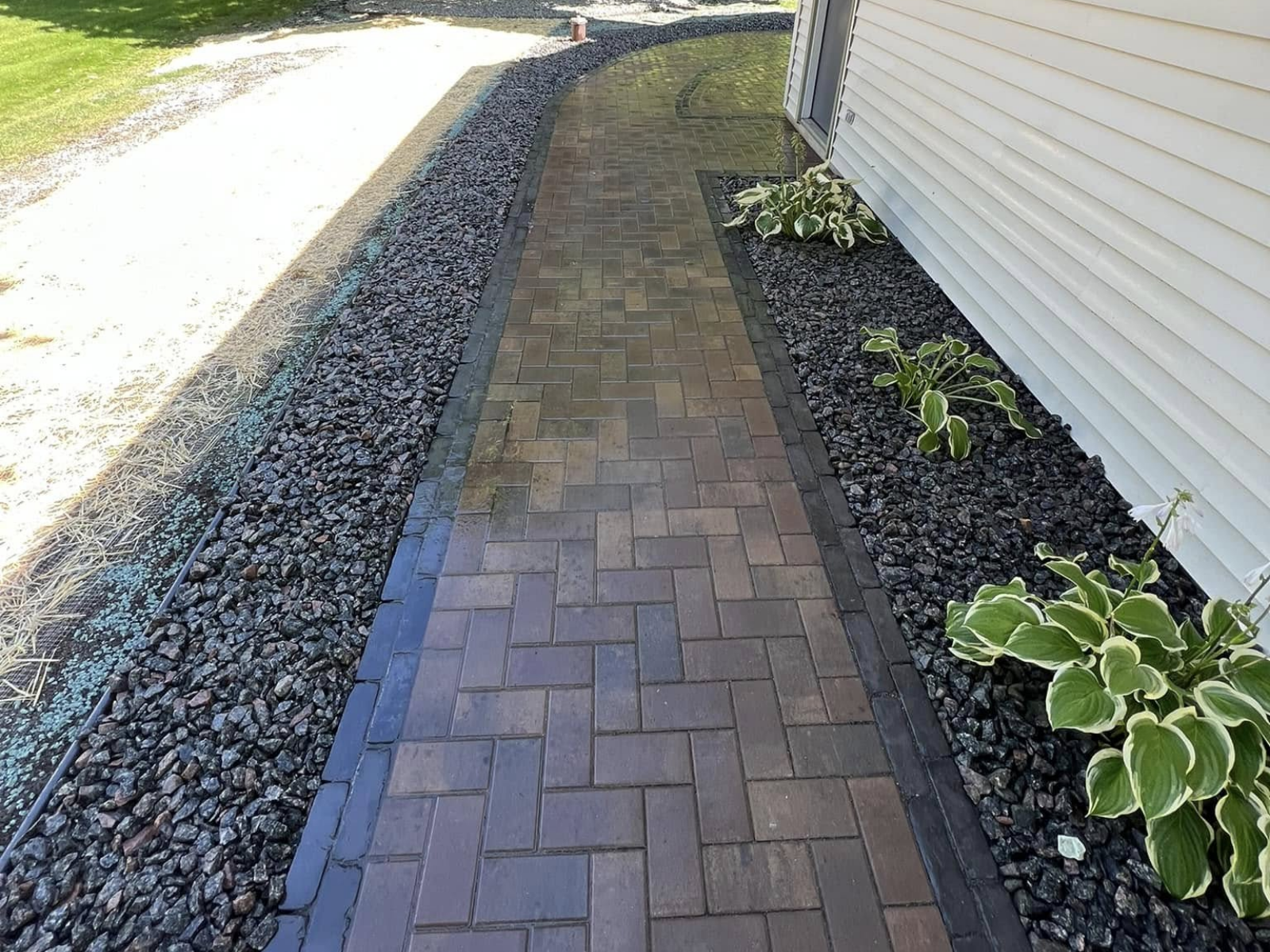 Brick walkway beside a house, bordered by black rock landscaping and green hostas.