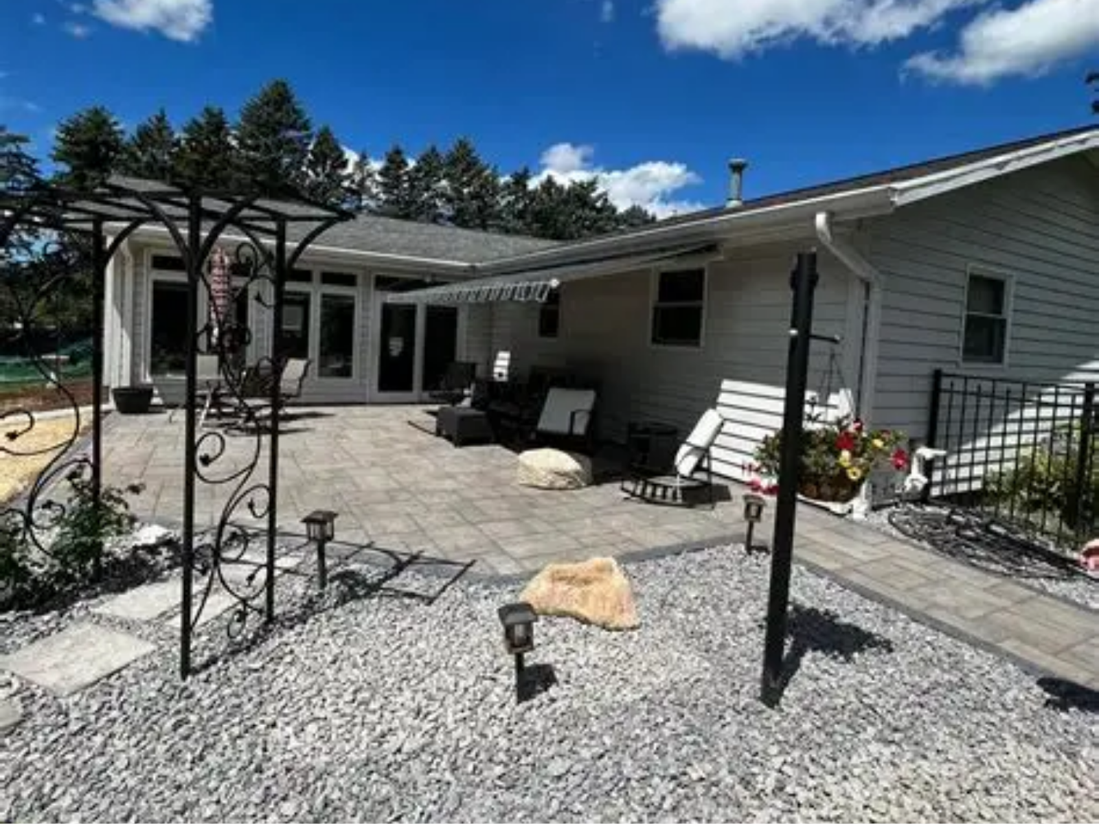 Back patio with gravel courtyard, covered seating area, and black metal garden trellis under a blue sky