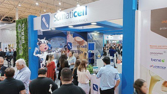Estande lotado em uma feira comercial, com sinalização azul e branca e participantes reunidos em torno de um expositor.