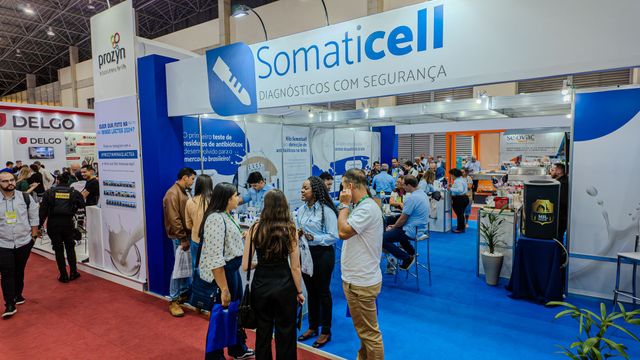Estande da “Somaticell” em uma feira comercial, com pessoas conversando sob placas azuis e brancas e um tapete vermelho.