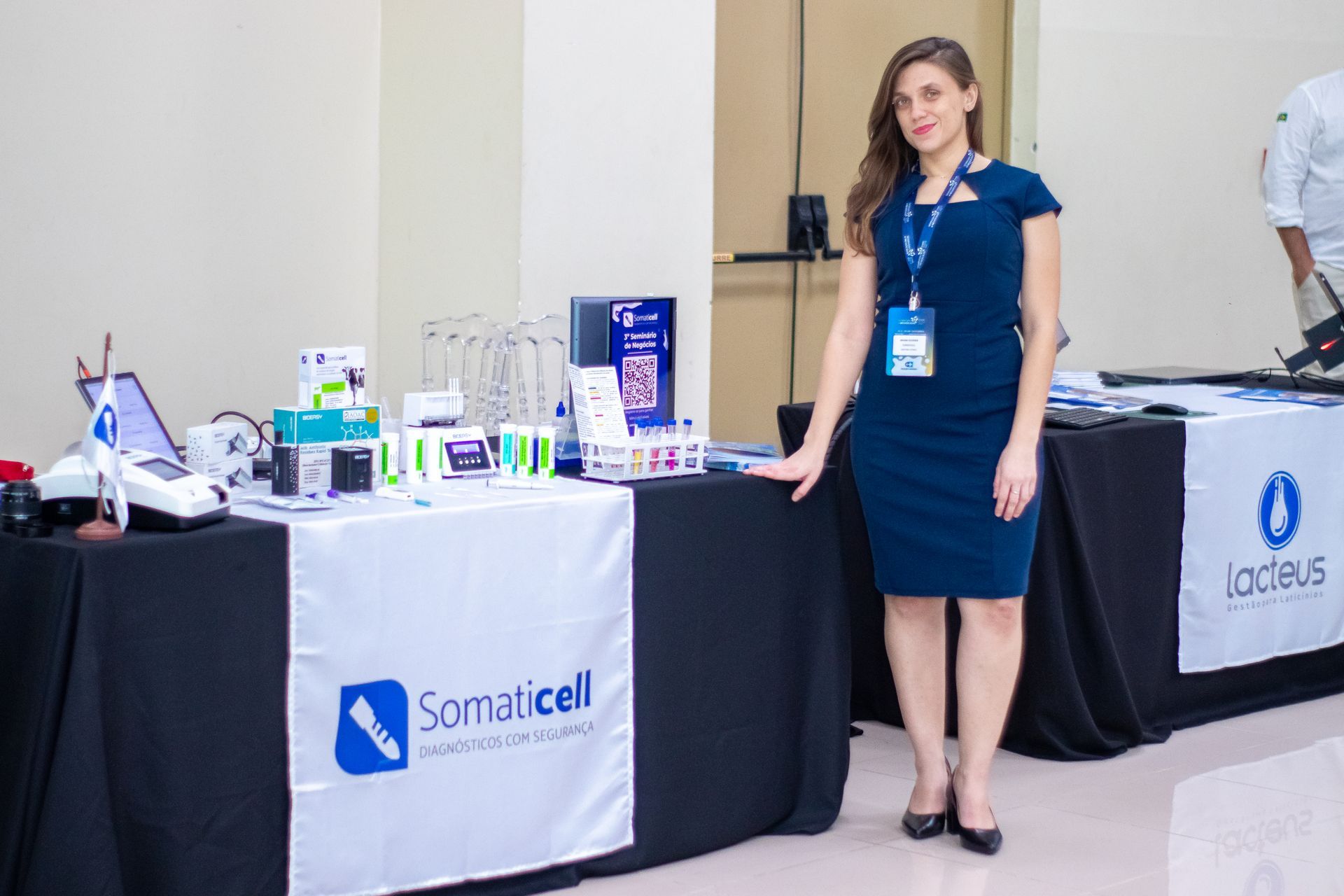 Uma mulher está ao lado de um estande de feira comercial com banners da Somacel e da Laduce e displays de produtos.