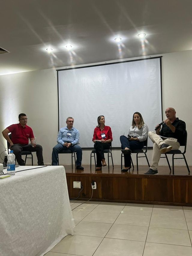 Mesa redonda com cinco palestrantes sentados em um pequeno palco em uma sala de conferências.