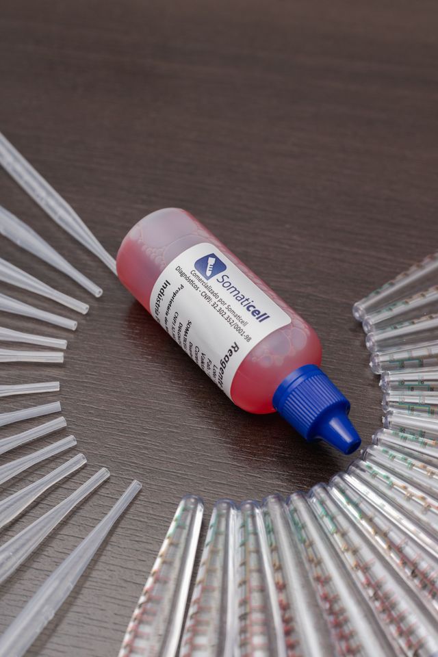 Frasco de apertar rosa com tampa azul, disposto entre hastes de plástico transparente sobre uma superfície marrom.