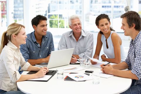 Team meeting in a bright office, with five people gathered around a laptop and documents on a table.