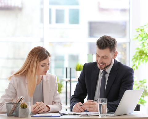 Two coworkers in business attire reviewing documents at a bright office desk with a laptop and coffee