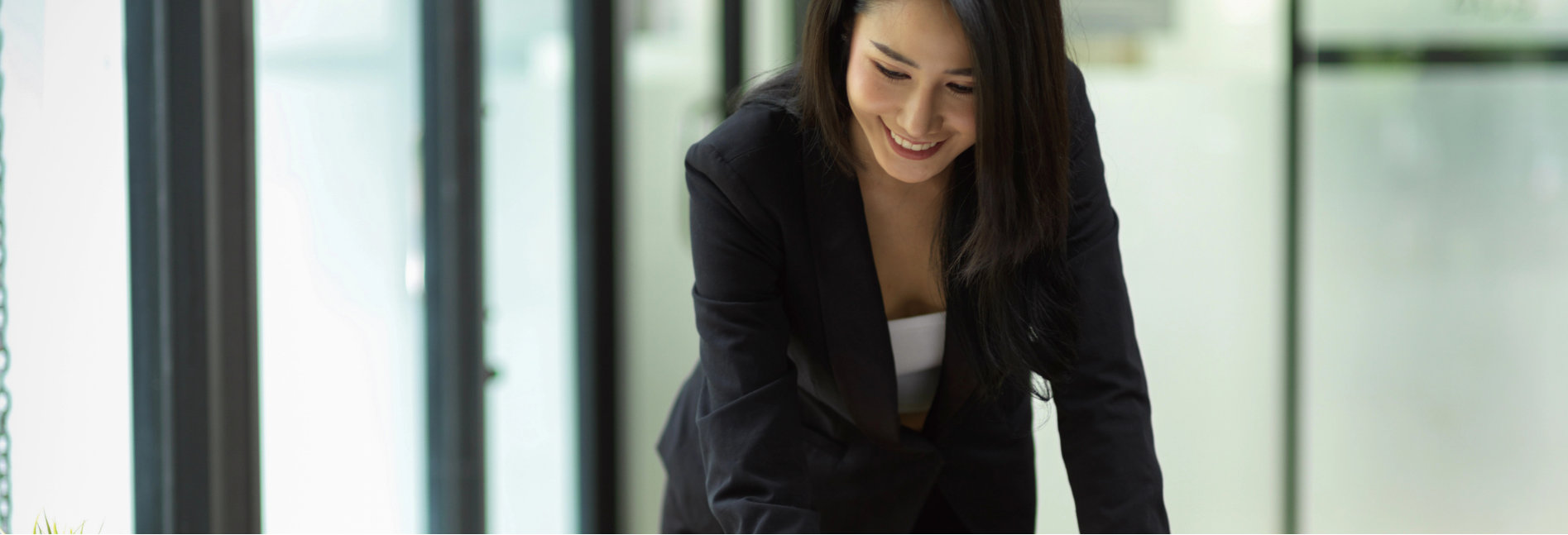 Woman in a dark blazer smiling and leaning over a desk in a modern office.
