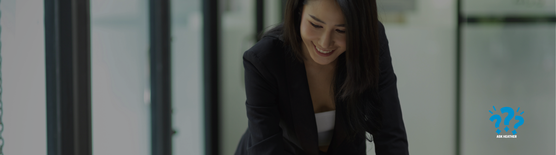 Woman in a dark blazer smiling and leaning over a desk in a modern office.