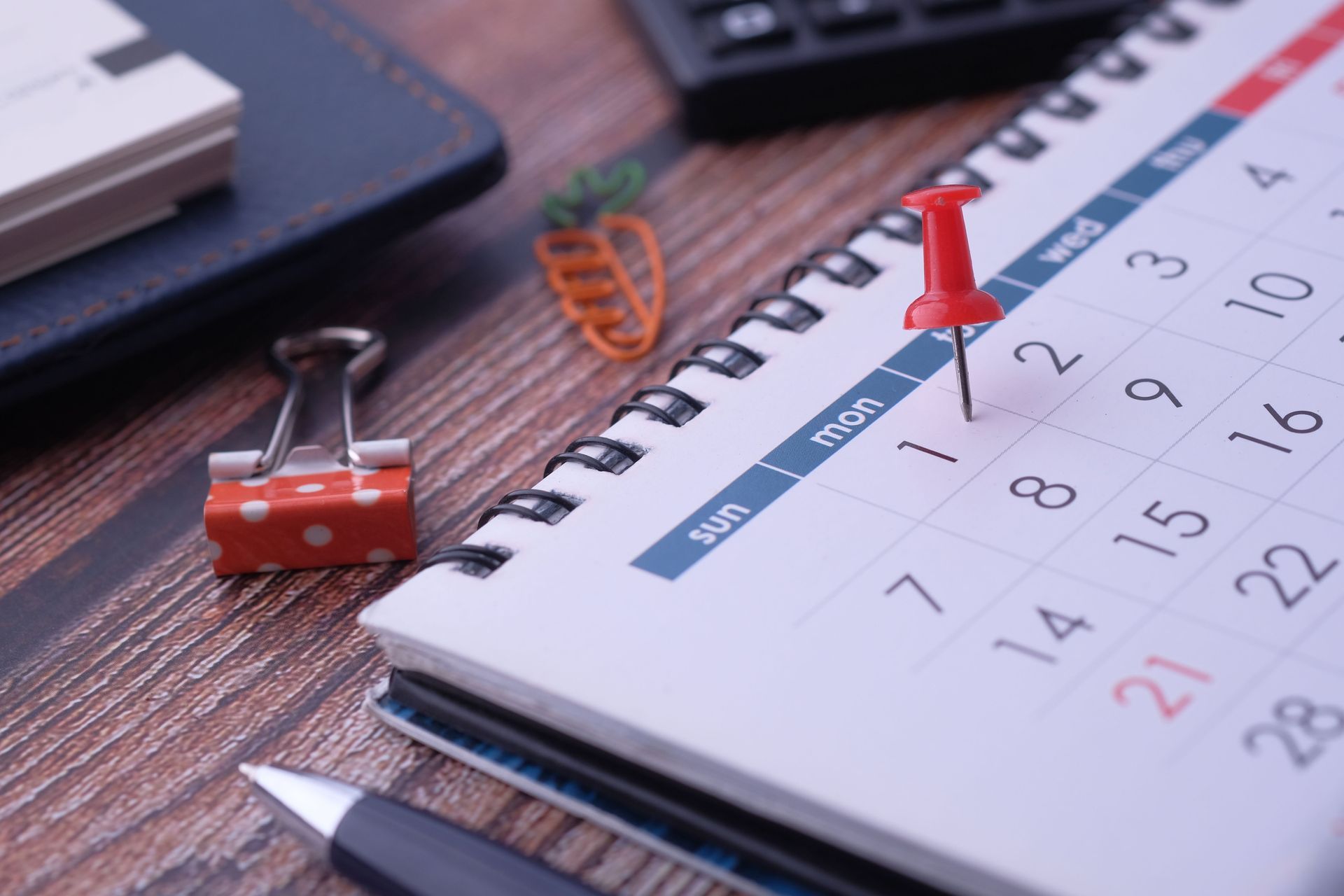 Desk calendar with a red pushpin, small red binder clip, and notebook on a wooden table