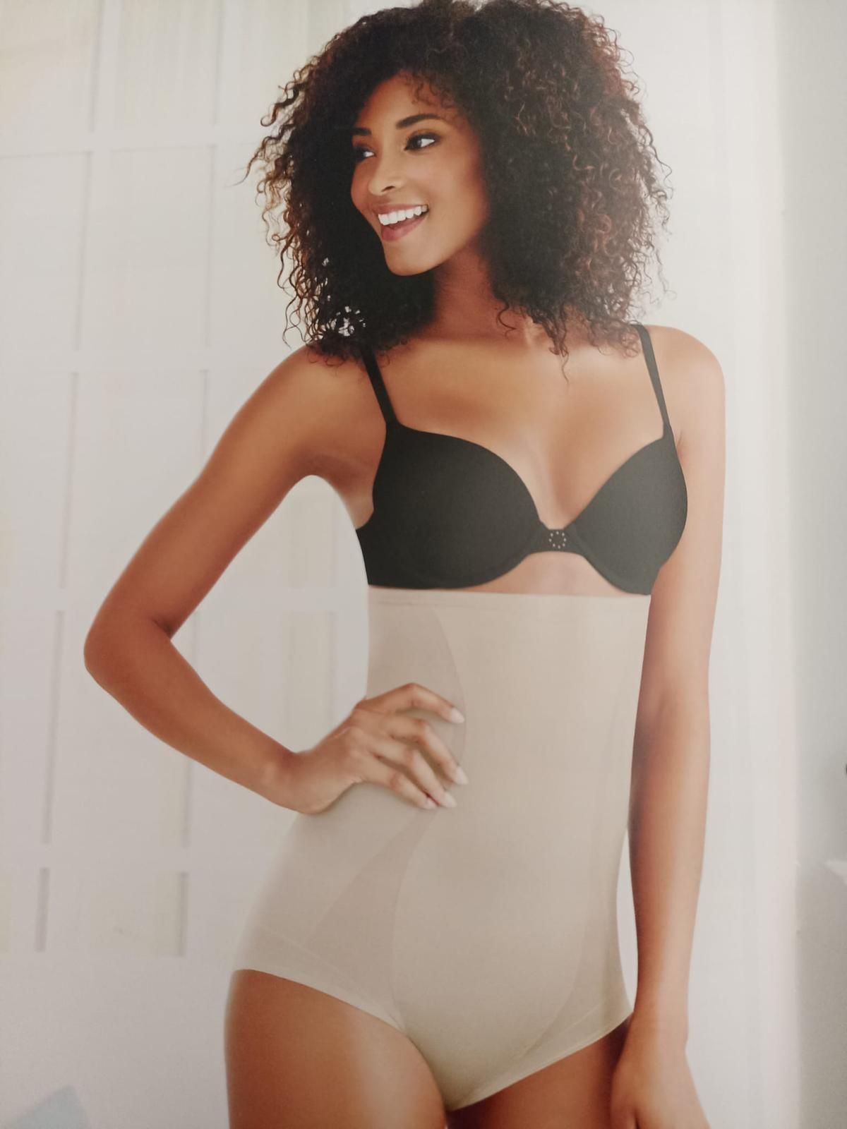 Donna in reggiseno nero e guaina beige a vita alta, sorridente e in posa.