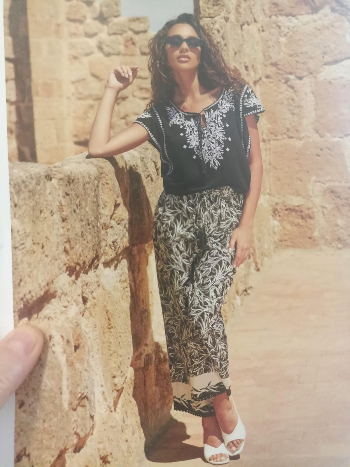 Donna con top nero ricamato e pantaloni fantasia, appoggiata a un muro di pietra, con occhiali da sole.