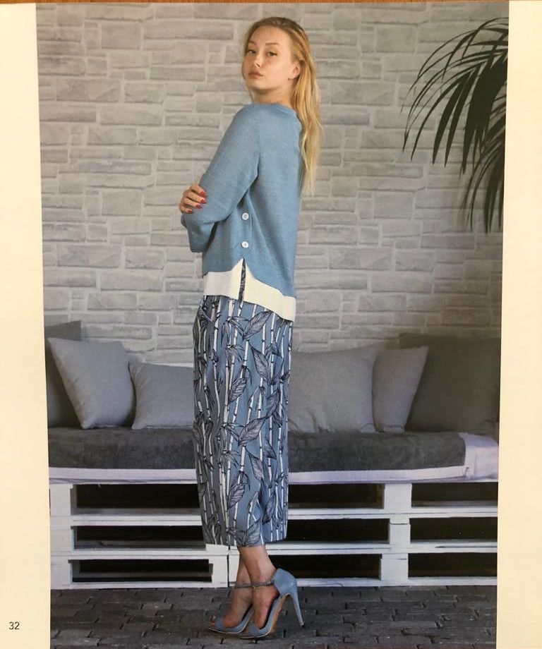Donna con maglione blu e pantaloni fantasia, in piedi accanto a un divano di pallet di fronte a un muro di mattoni.