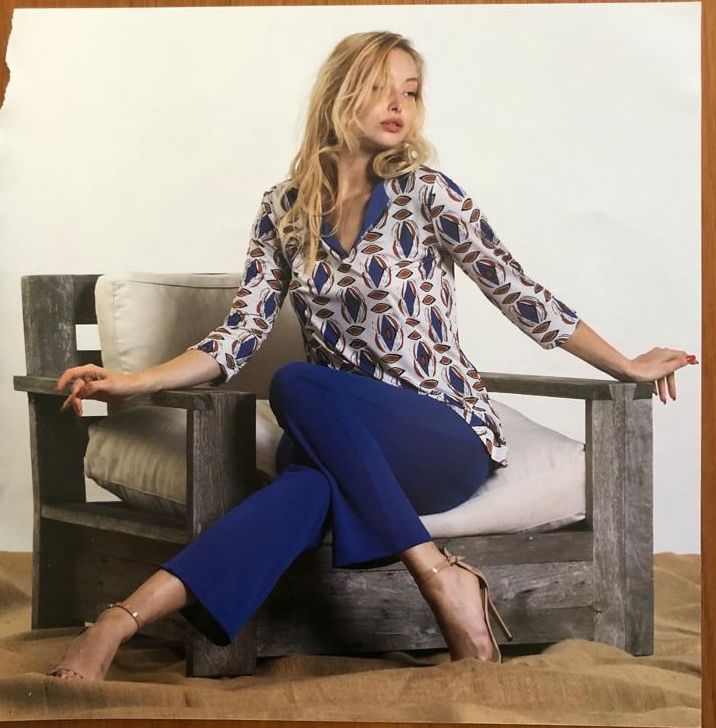 Una donna con una maglietta fantasia e pantaloni blu è seduta su una sedia di legno, con le gambe incrociate, e sorride.