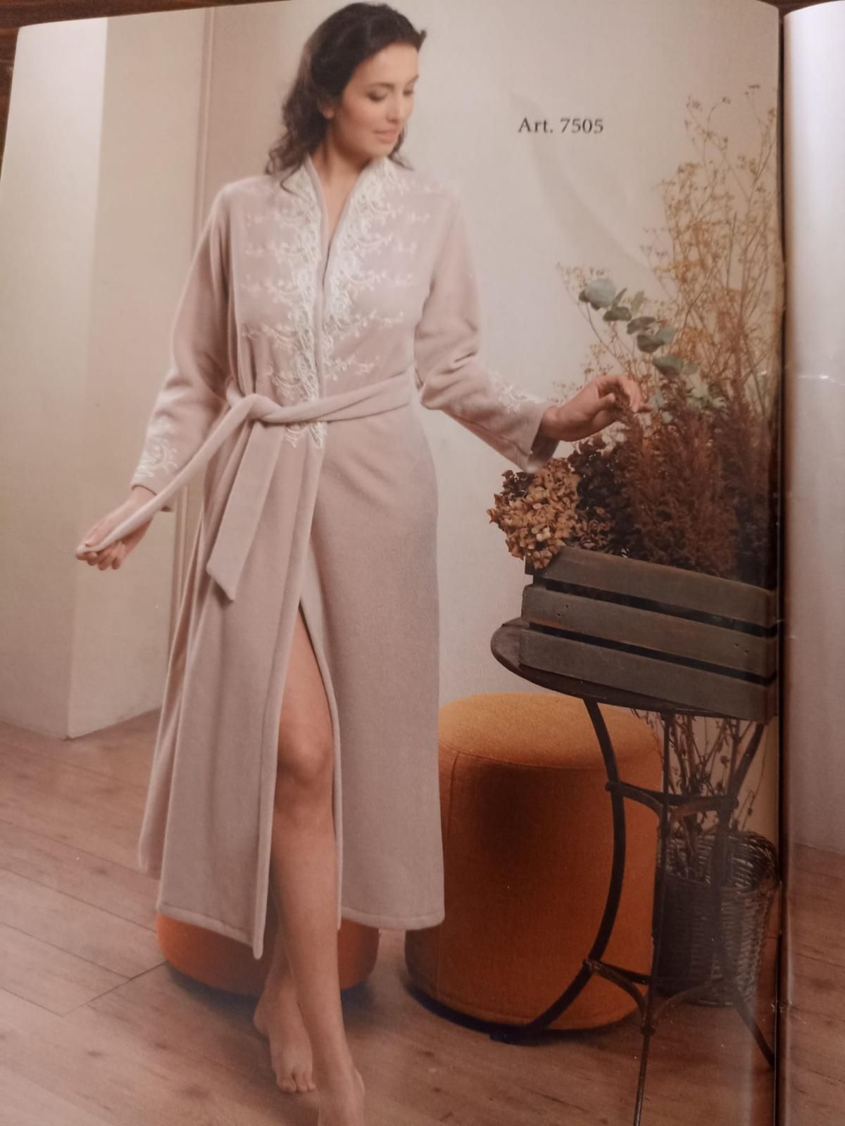 Donna con una veste beige chiaro con ricami floreali, in piedi accanto a un tavolo con fiori secchi.