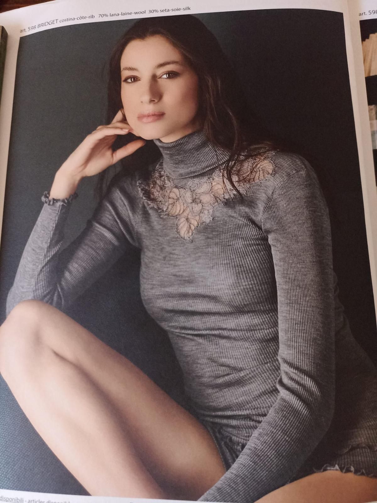 Donna con abito grigio a collo alto, appoggiata sul gomito, sorridente leggermente, su uno sfondo scuro.