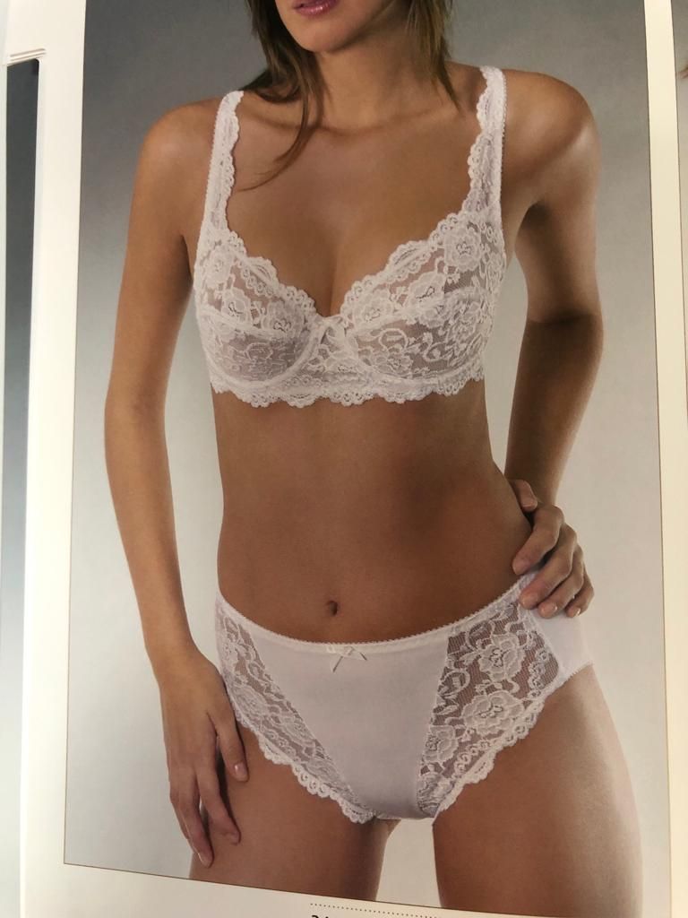 Donna con reggiseno di pizzo bianco e biancheria intima coordinata. È in piedi, in posa su uno sfondo grigio chiaro.