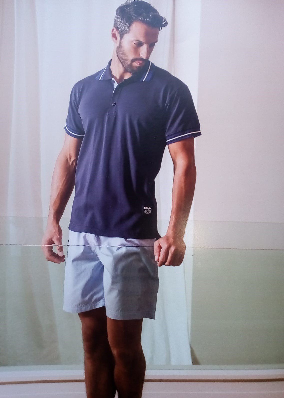 Un uomo con una polo blu navy e pantaloncini azzurri è in piedi, rivolto in avanti, su uno sfondo bianco e verde.