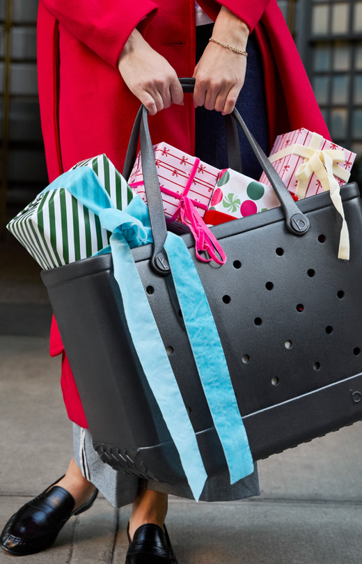 Woman in red coat holding a large black tote bag filled with wrapped gifts.