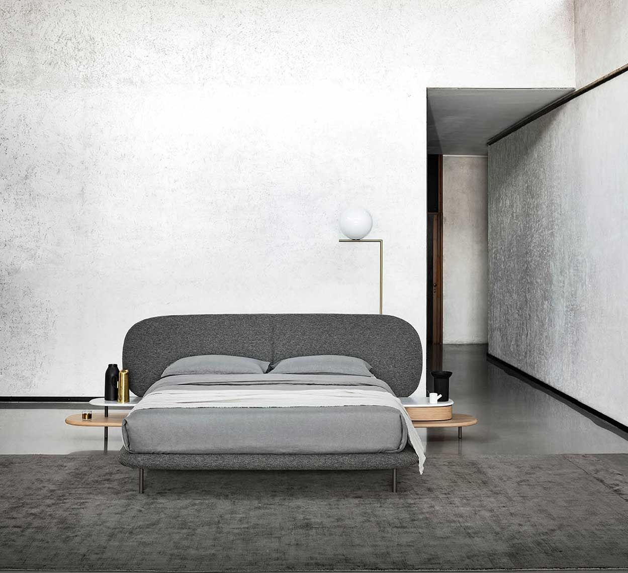Camera da letto minimalista con letto grigio, pareti bianche e una moderna lampada da terra con luce soffusa.