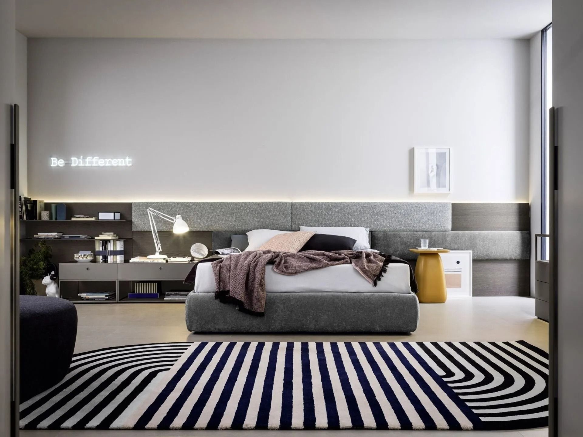 Camera da letto minimalista con letto grigio, tappeto a righe e mensole a muro in un moderno arredamento neutro.