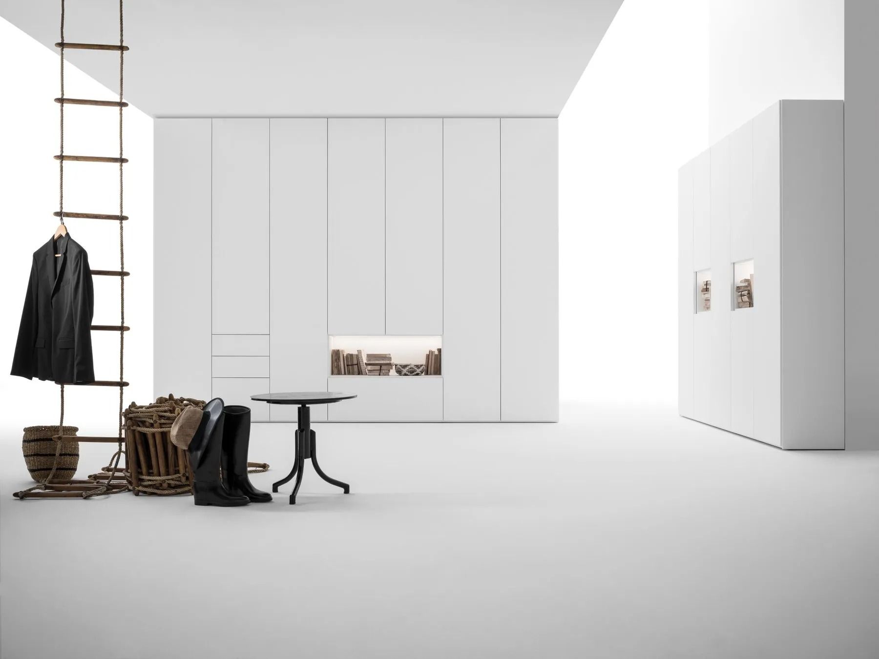 Stanza minimalista bianca con scala, cappotto nero appeso, stivali, tavolo e armadi a muro.