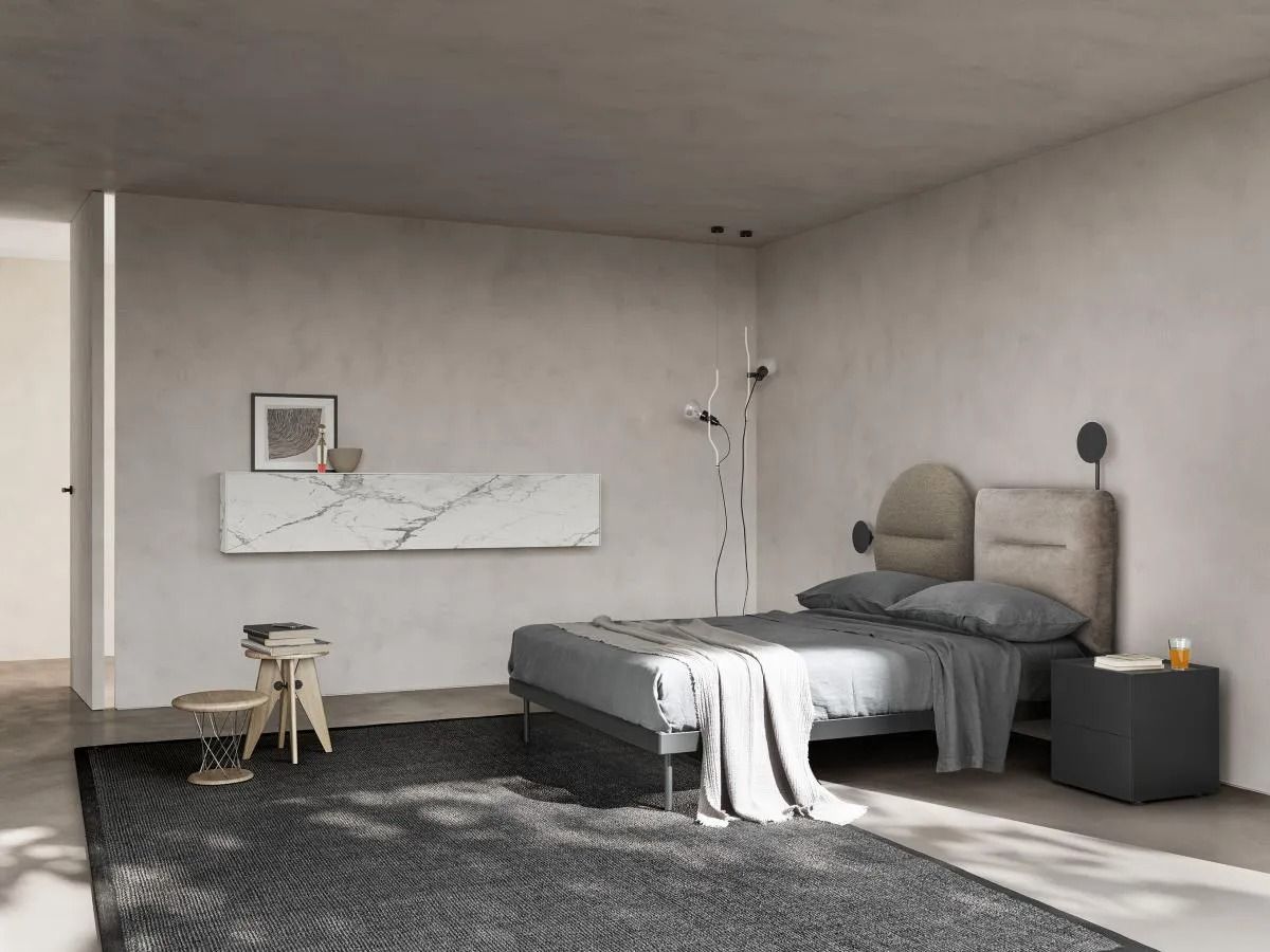 Camera da letto minimalista con letto grigio, pareti bianche, tappeto scuro e un piccolo sgabello vicino alla finestra.