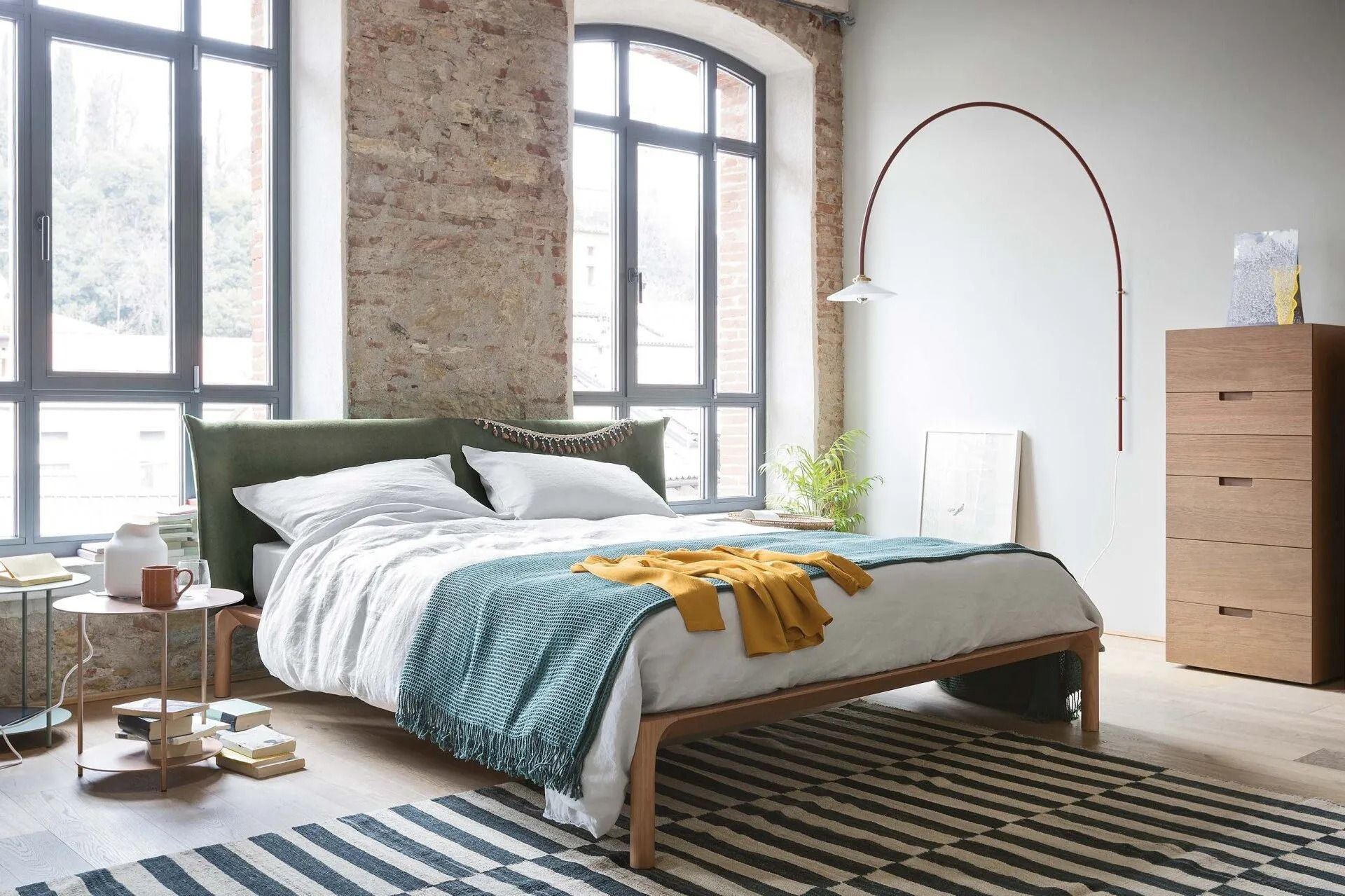 Camera da letto luminosa con ampie finestre, letto con lenzuola blu e gialle, tappeto a righe e comò in legno.
