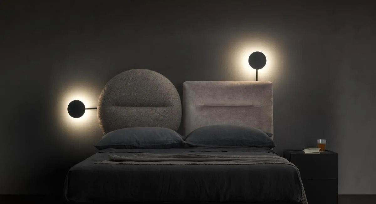 Camera da letto moderna con letto scuro, testiera rotonda imbottita e due lampade da parete luminose su una parete grigia.