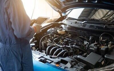 Una persona inspecciona el motor abierto de un coche en un garaje, con luz solar y herramientas cerca.