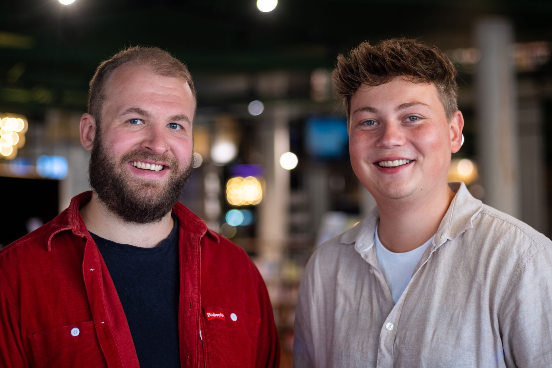 Twee lachende mannen staan binnen; de ene in een rode jas, de andere in een licht overhemd, met een wazige achtergrond.