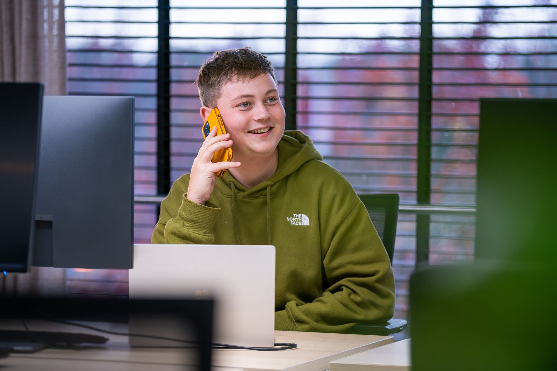 Persoon in groene hoodie, pratend aan een gele telefoon, glimlachend, zittend aan een bureau met een laptop en computermonitoren.