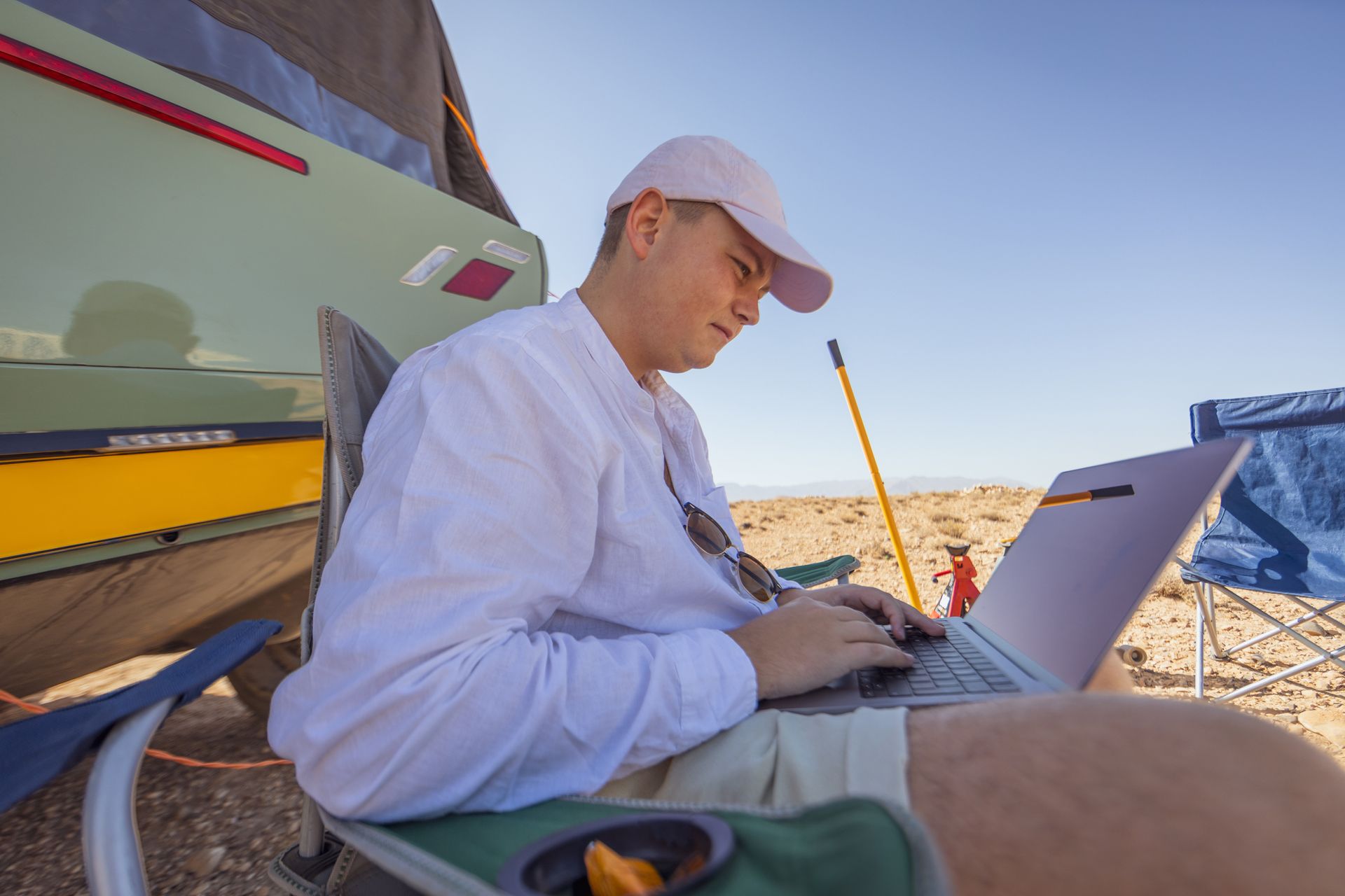 Persoon in wit shirt en hoed typt op een laptop tijdens een kampeertrip in een zonnige woestijn.