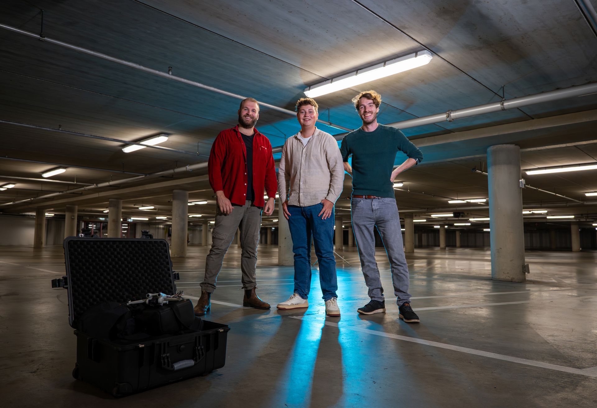 Drie mensen staan in een parkeergarage; één van hen bij een open koffer met apparatuur. Donkere, holle ruimte met plafondverlichting.