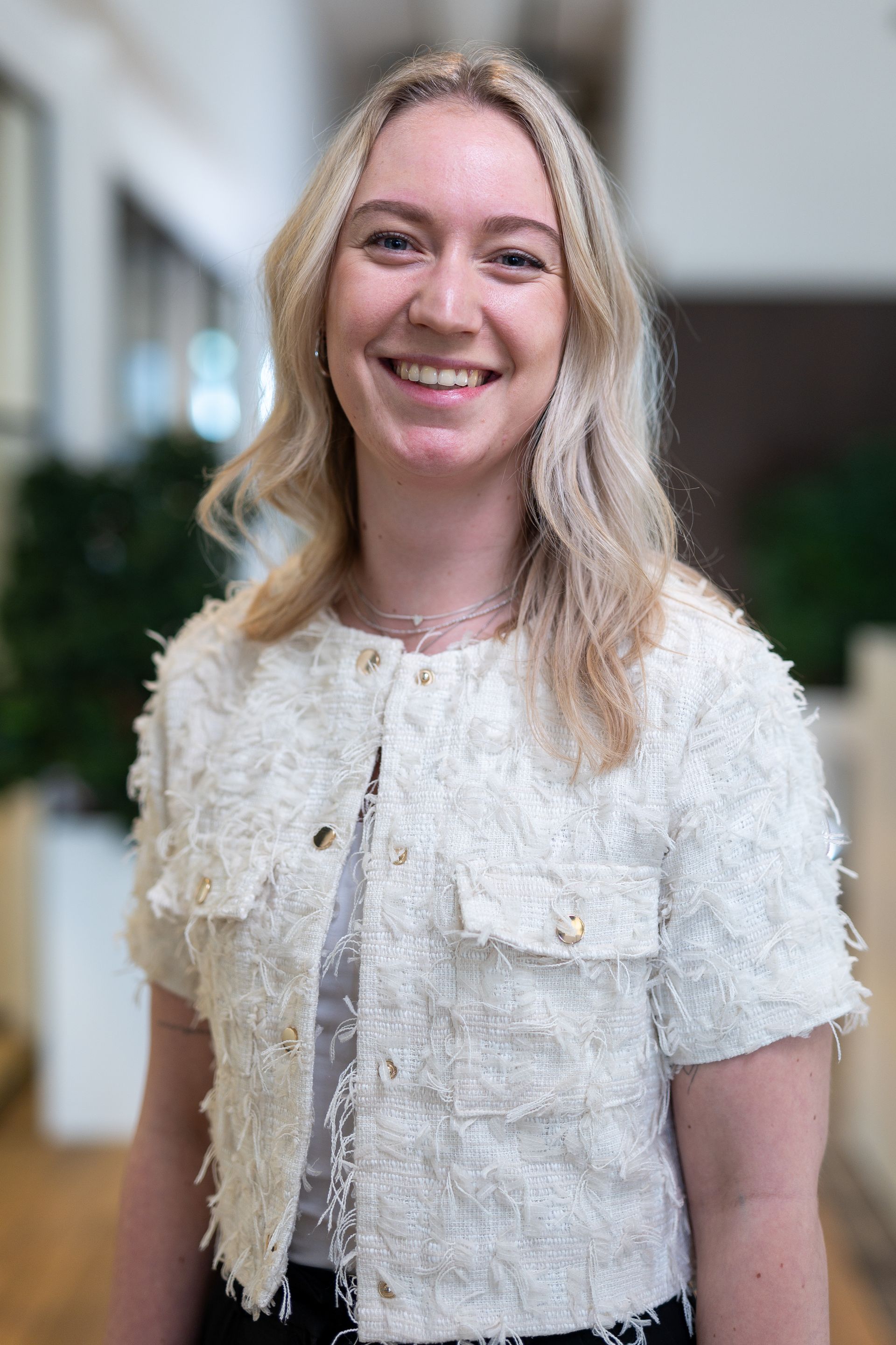 Blonde vrouw lacht, draagt ​​een witte jas met textuur, en staat binnen.