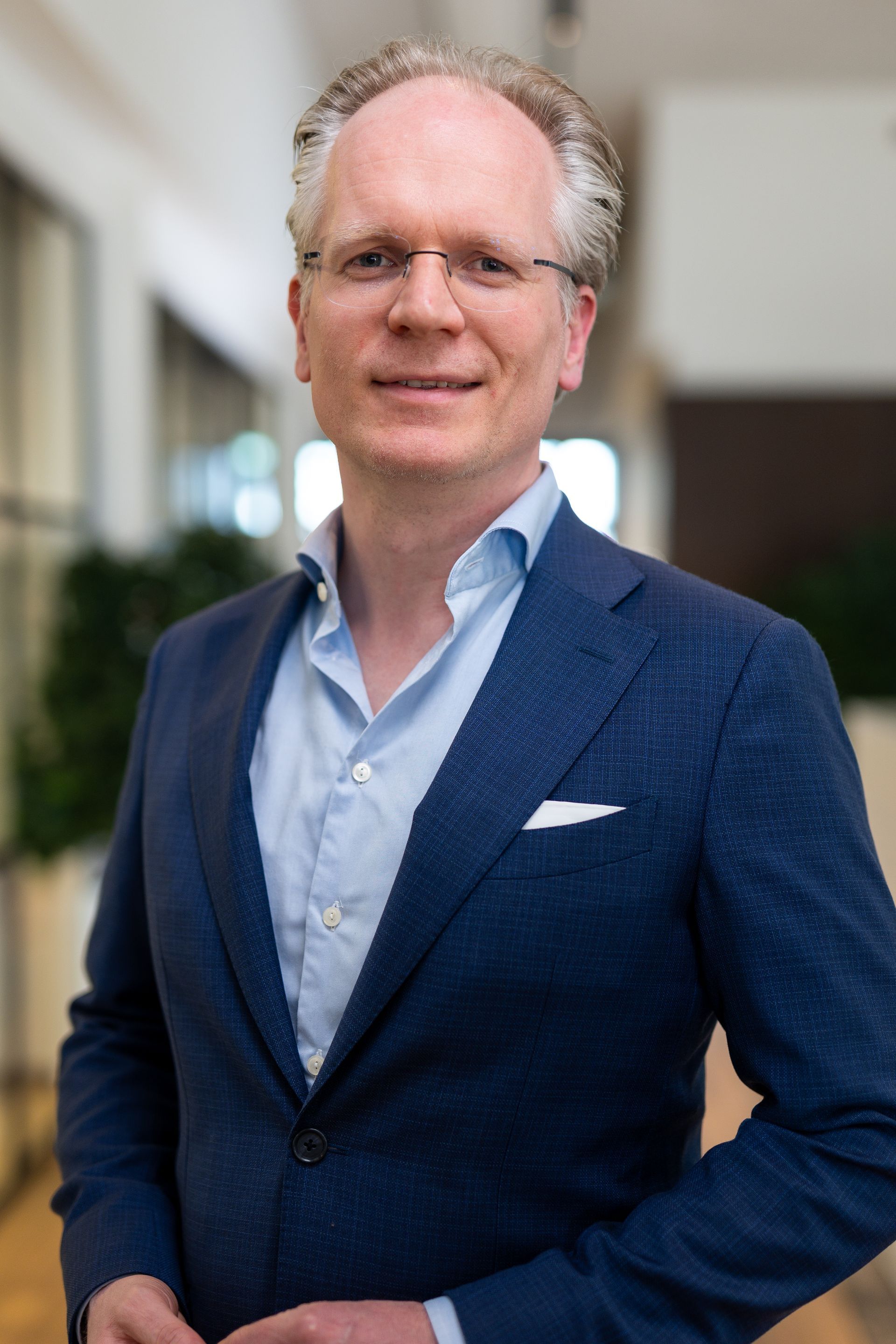 Een man in een blauwe blazer en een lichtblauw overhemd staat lachend in een kantoorgang.