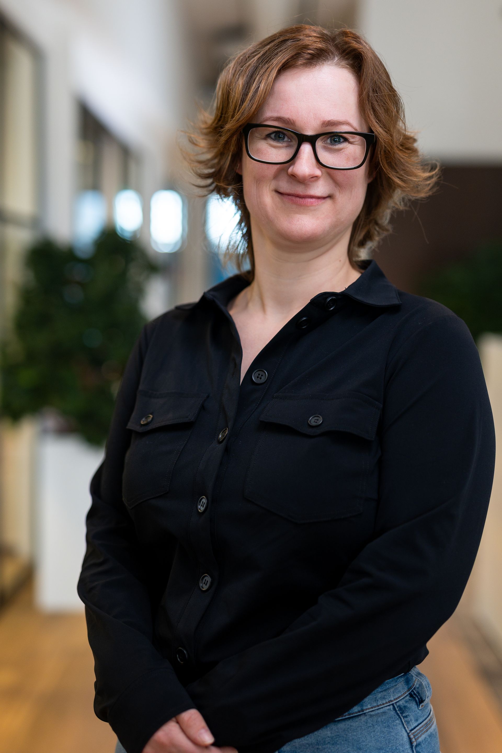 Vrouw met bril en zwarte blouse, lachend in een kantooromgeving.