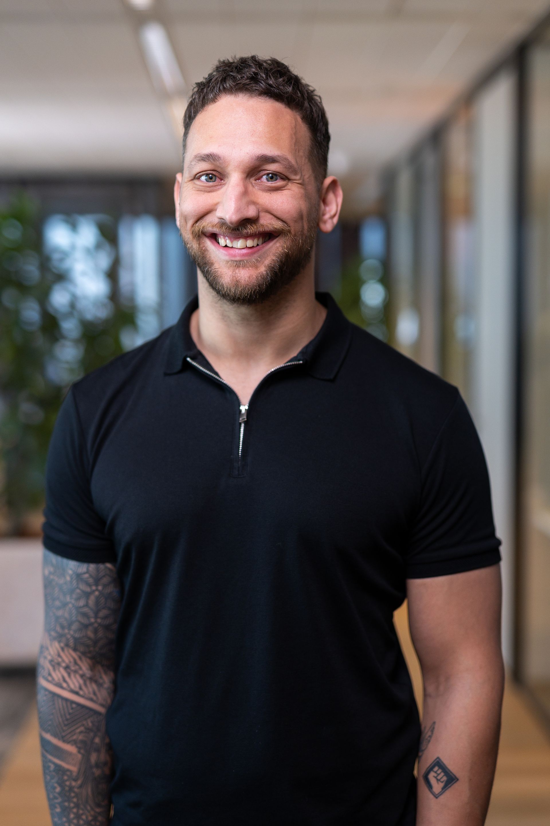 Man met kort bruin krullend haar en een baard, lachend, gekleed in een zwart poloshirt, met een getatoeëerde arm, in een kantooromgeving.