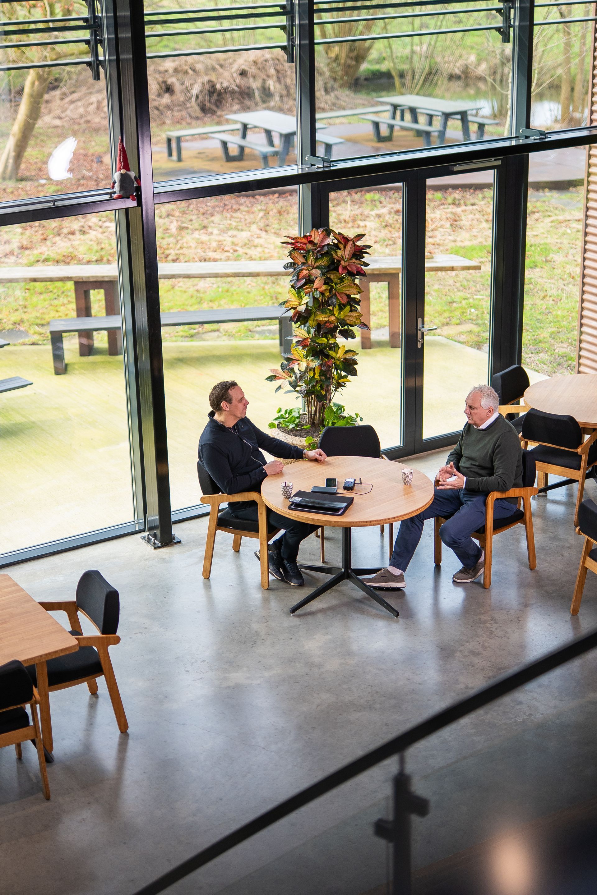 Twee mannen zitten aan een ronde tafel binnen; grote ramen bieden uitzicht op het terras.
