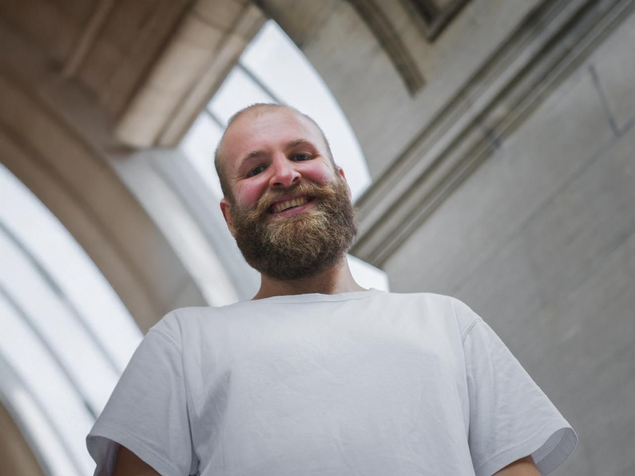 Een man met een volle baard, gekleed in een wit T-shirt, staat lachend binnen onder een gebogen plafond.