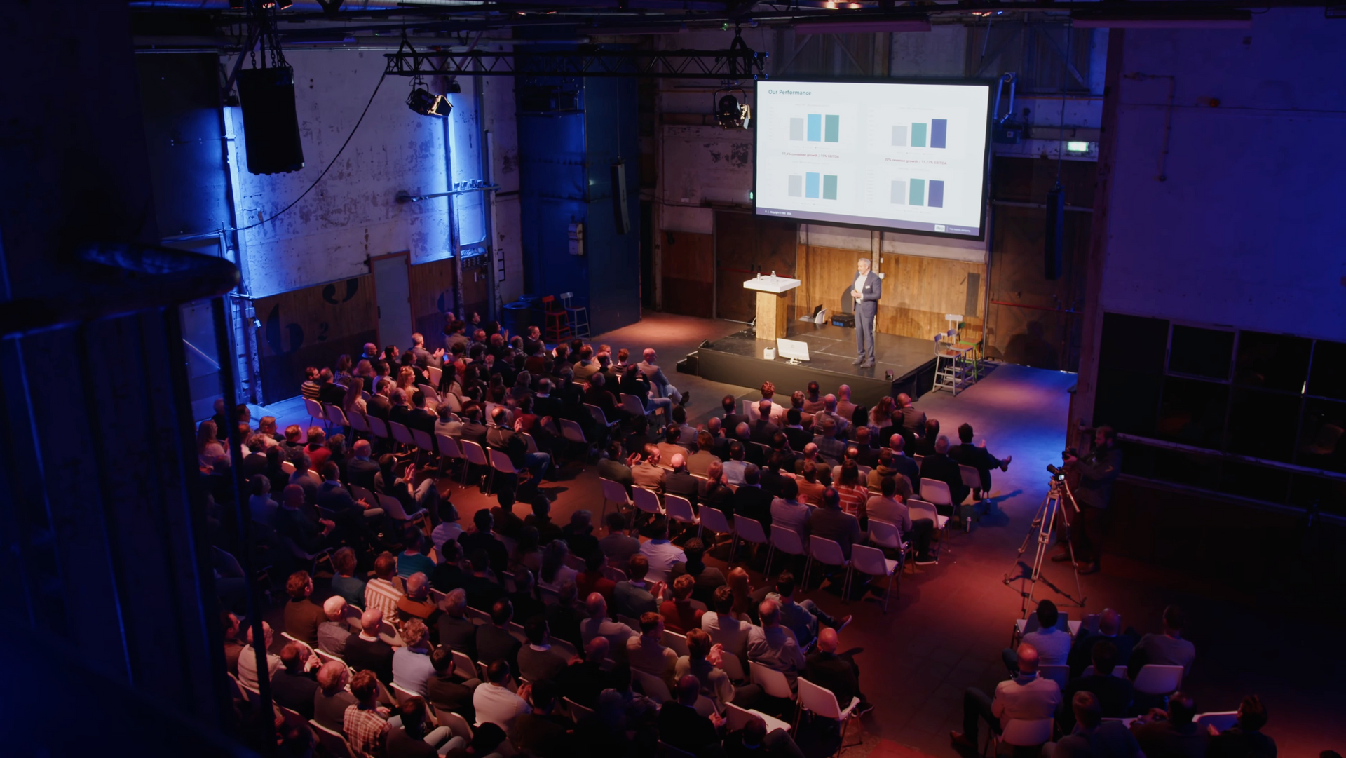Een spreker geeft een presentatie op een podium voor een publiek dat in rijen zit; op een scherm wordt een presentatie met kleurstalen weergegeven.