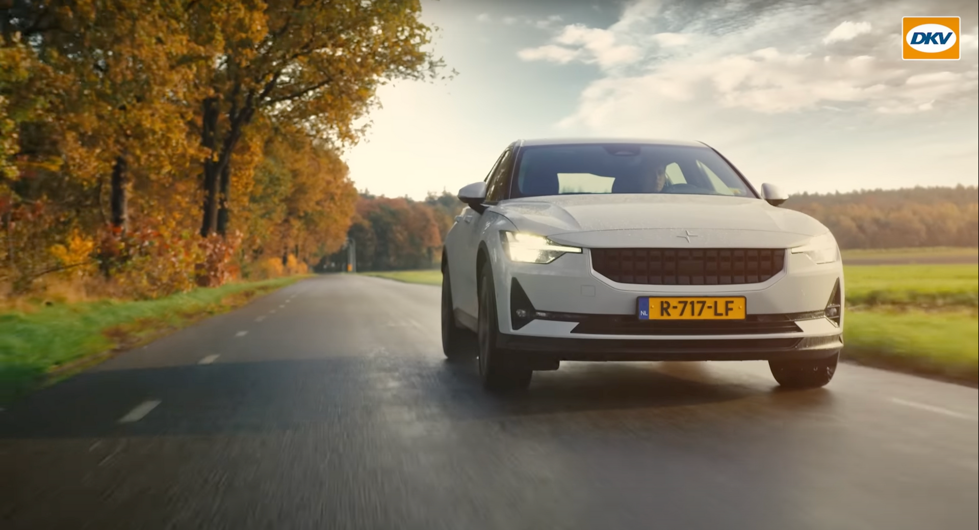 Een witte auto rijdt over een natte weg in de herfst. Het zonlicht filtert door de bomen.