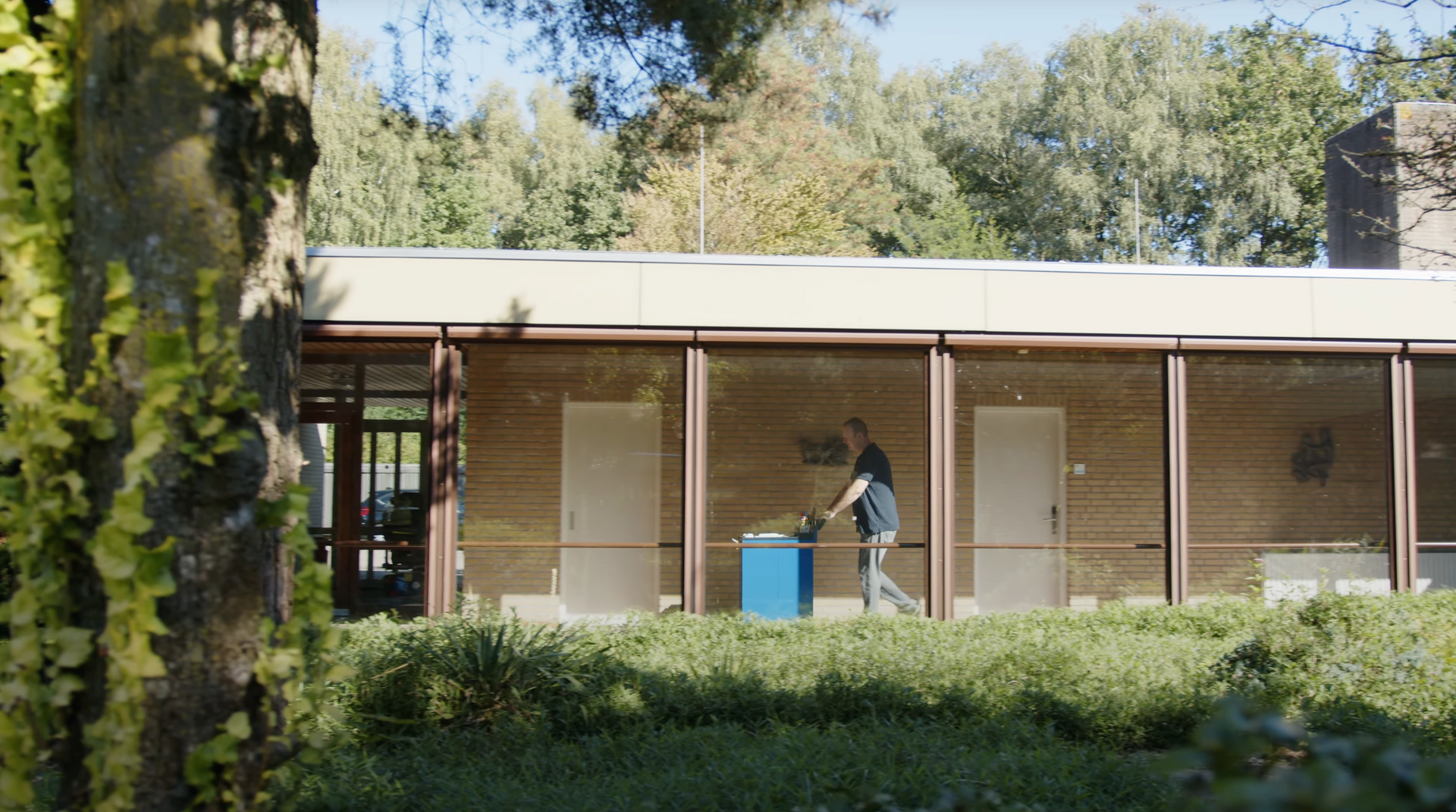 Een persoon duwt een blauwe kar in een gebouw met een groot raam. Het gebouw heeft een plat dak en op de achtergrond zijn bomen te zien.