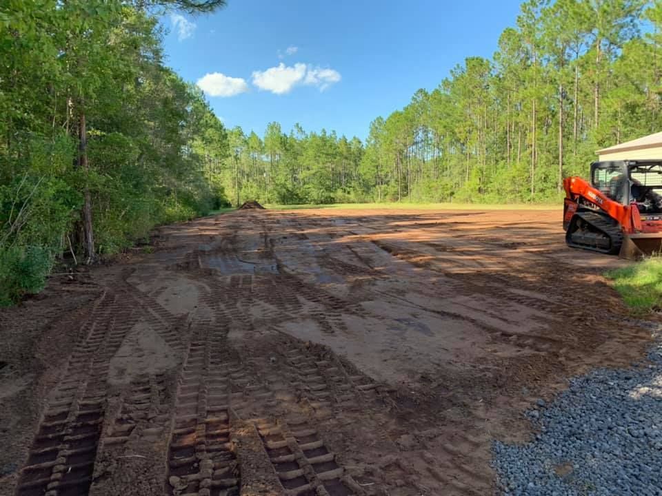 Cleared dirt field with track marks, surrounded by trees, with an orange skid steer on the right.
