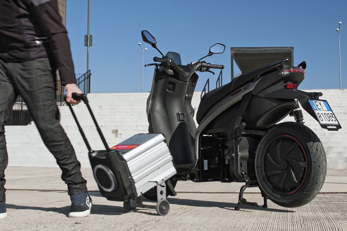 Un uomo spinge una valigia accanto a uno scooter.
