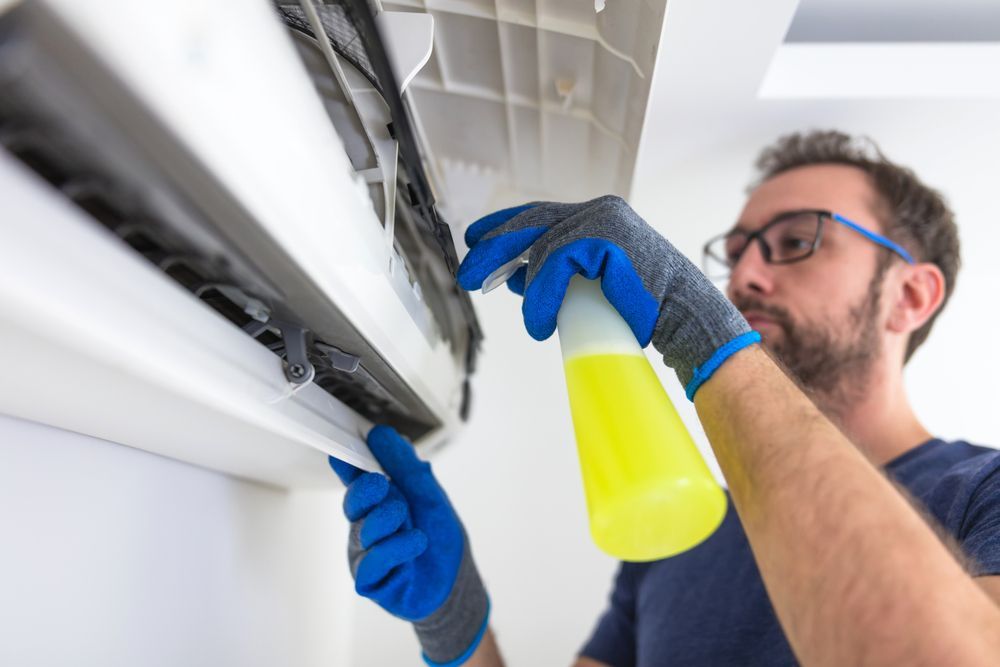 A Man is Cleaning an Air Conditioner With a Spray Bottle — Revive Air Conditioning in Oran Park, NSW