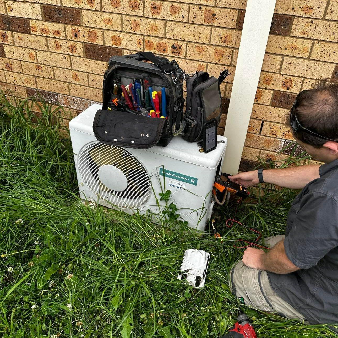 A Man Fixing an Air Conditioner on A Brick Wall — Revive Air Conditioning in Campbelltown, NSW