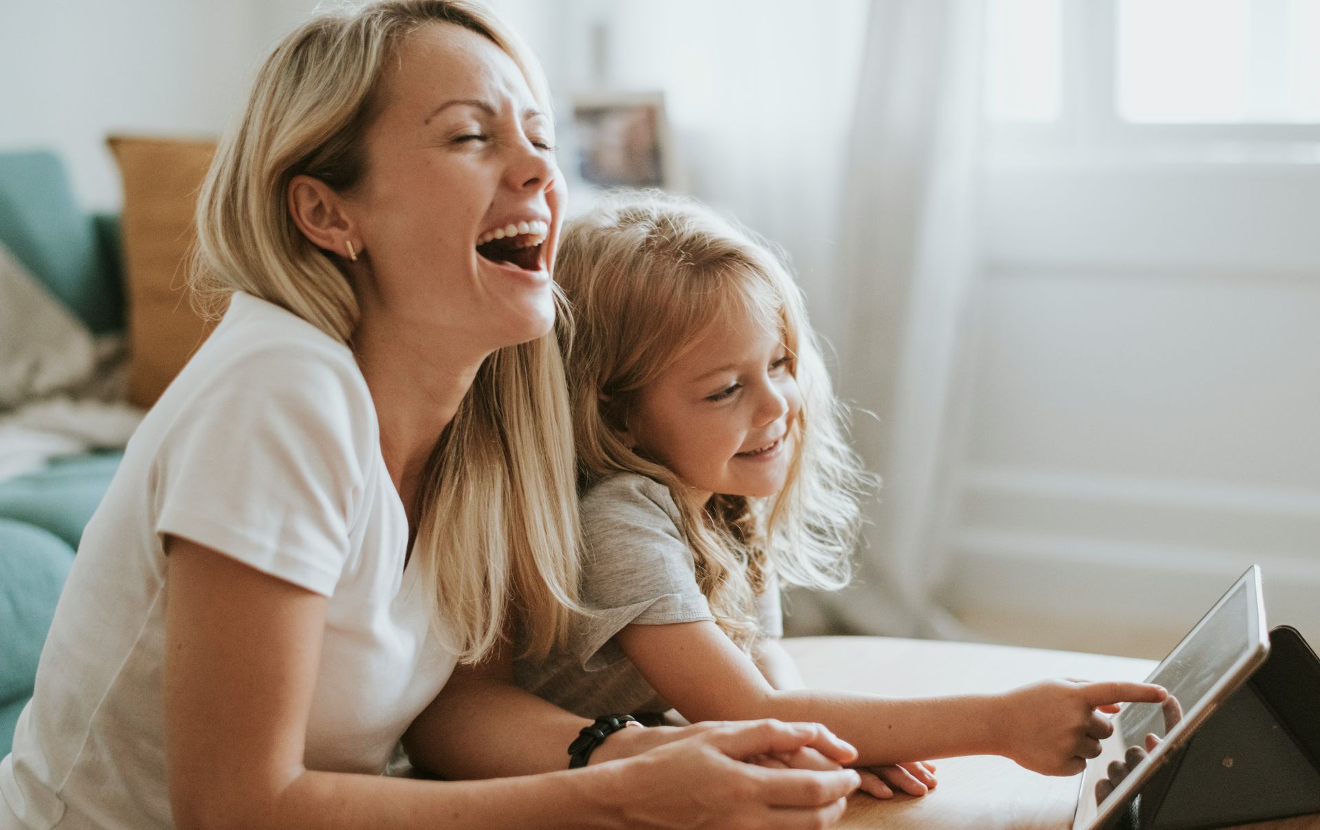 Woman and child laughing while using a laptop on a couch in a bright room