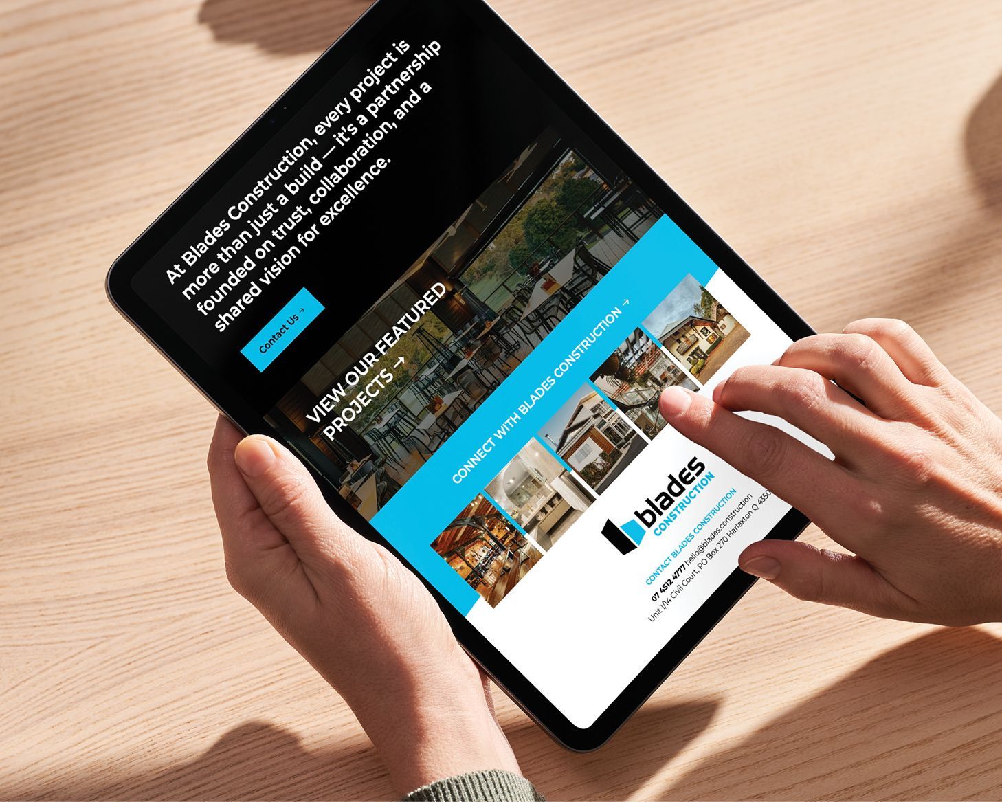 Hands using a tablet on a wooden table, showing a website with a blue banner and small photo thumbnails.