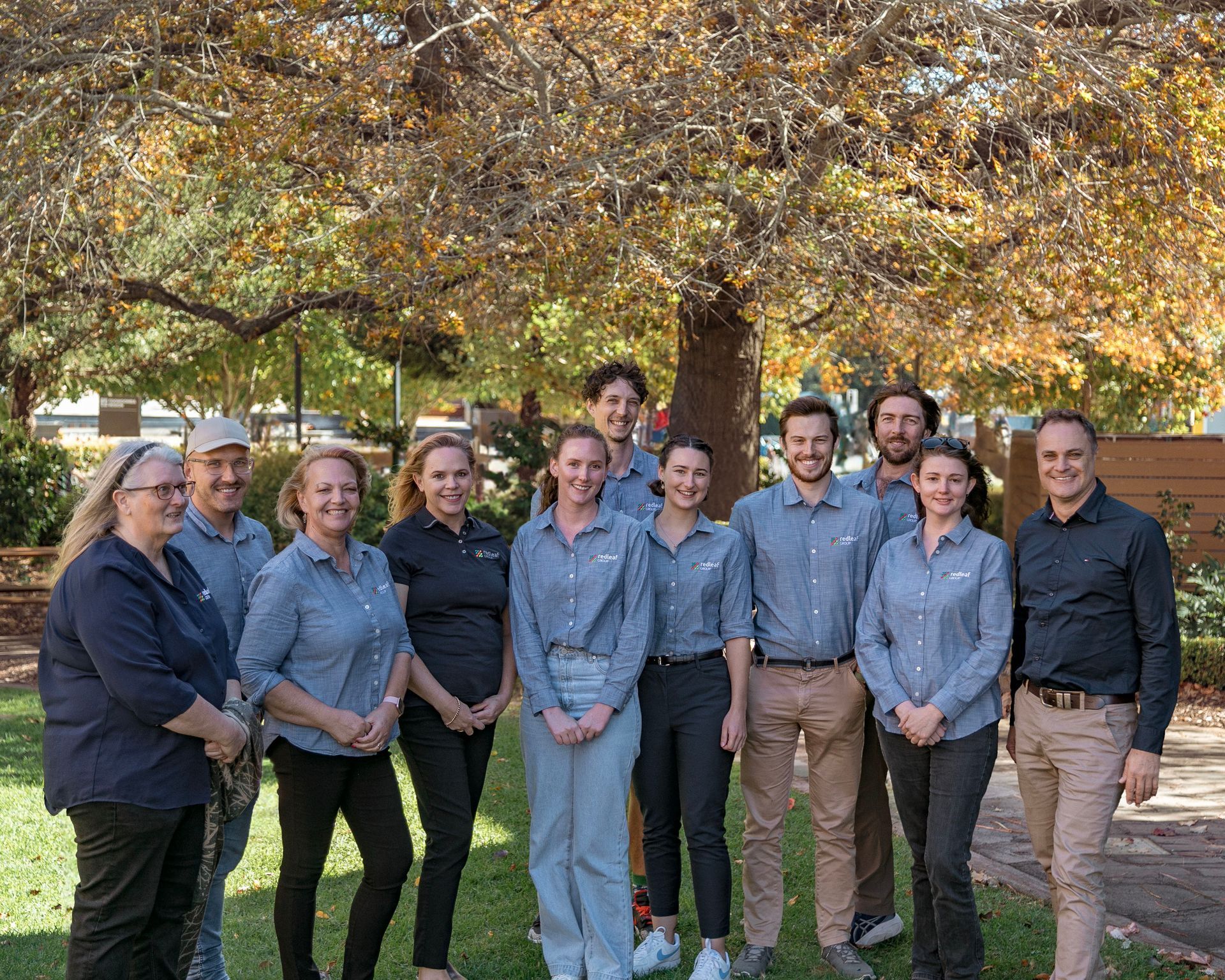 Group of people standing outdoors beneath autumn trees, wearing matching blue shirts and smiling at the camera