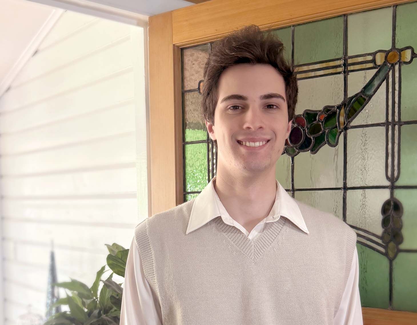 Person smiling indoors beside a stained-glass window, wearing a light collared sweater.