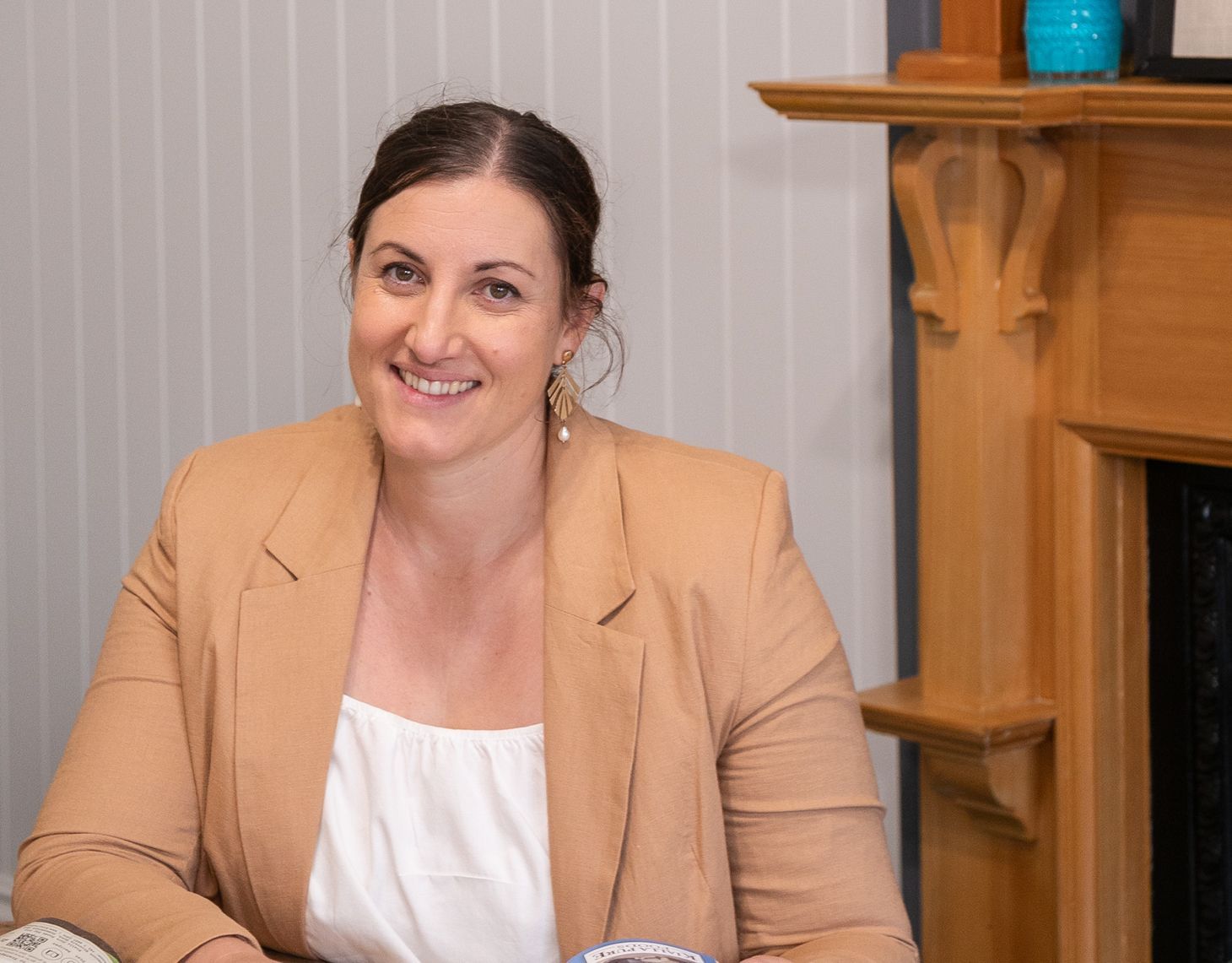 Smiling woman in a tan blazer seated indoors beside a wooden fireplace mantel